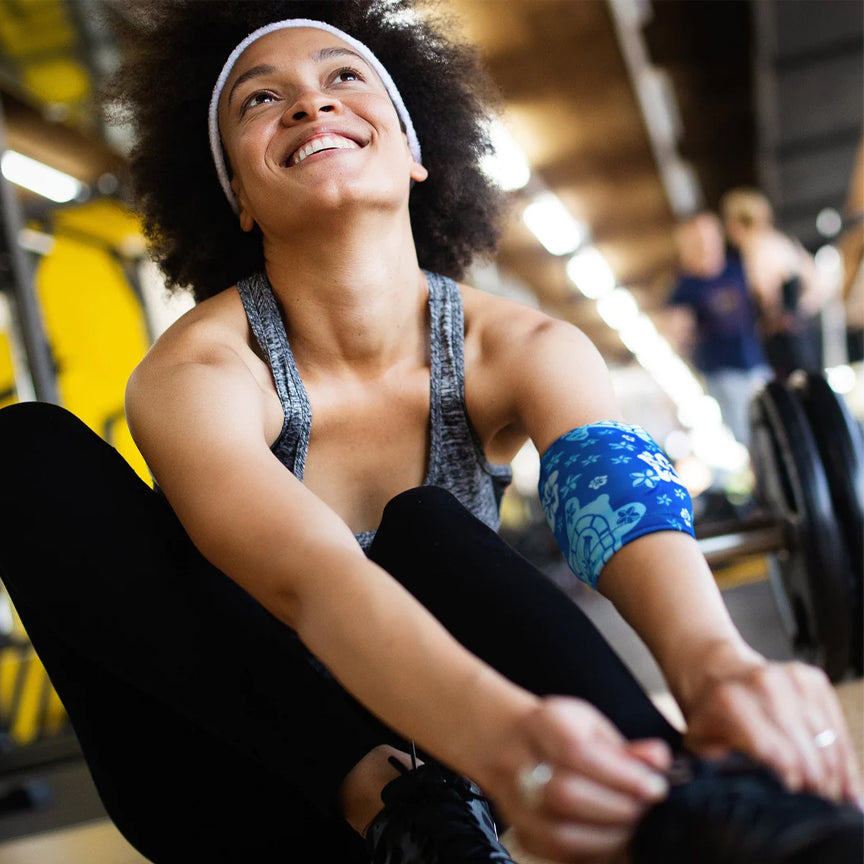 A woman in athletic wear is smiling while stretching on a yoga mat and wearing  blue HurtSkurt cold pack for workout recovery.