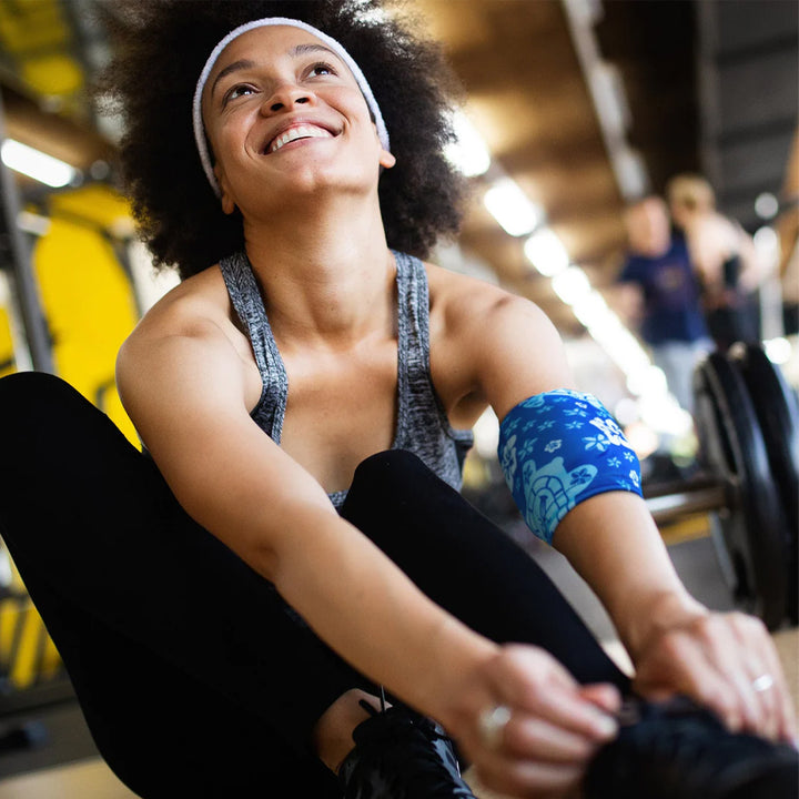 A woman in athletic wear is smiling while stretching on a yoga mat and wearing  blue HurtSkurt cold pack for workout recovery.